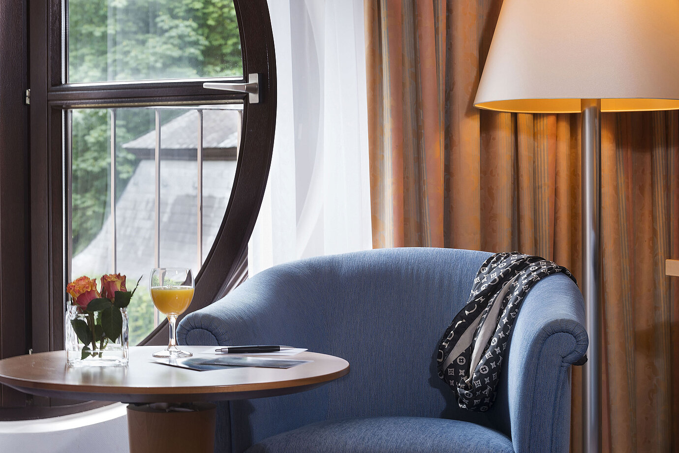 Accogliente angolo relax nella Camera Classic del Maritim Fulda con poltrona blu, finestra rotonda, fiori e succo d’arancia.