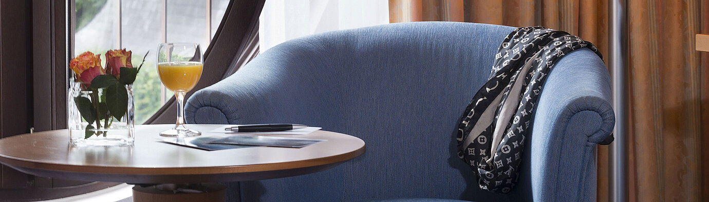 Cozy seating area in the Classic Room of Maritim Hotel Fulda with blue armchair, round window, flowers, and orange juice.