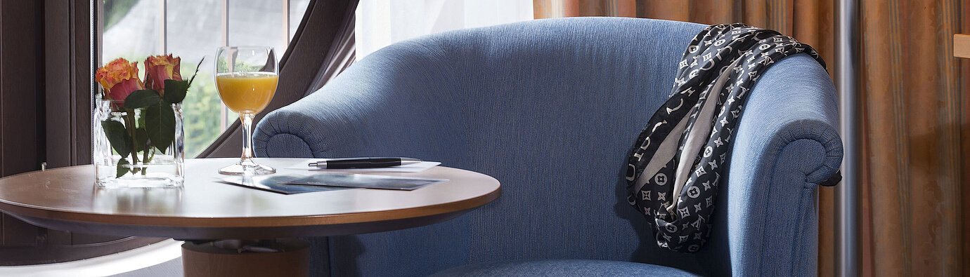Cozy seating area in the Classic Room of Maritim Hotel Fulda with blue armchair, round window, flowers, and orange juice.