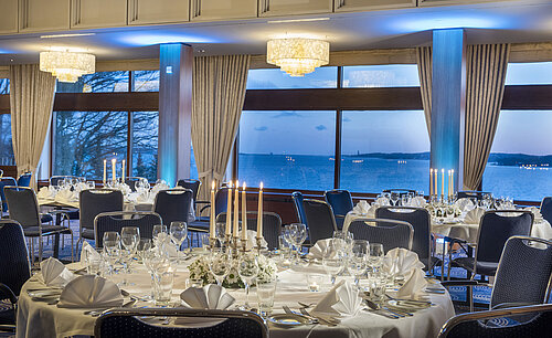 Maritim Hall in the Maritim Hotel Kiel, festively laid tables with candlelight, large windows and sea views at dusk.