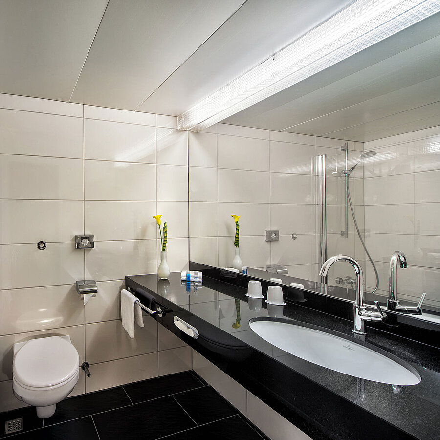Bagno del Maritim Hotel Kiel con vasca da bagno, schermo doccia in vetro, lavabo e grande specchio su una mensola nera.
