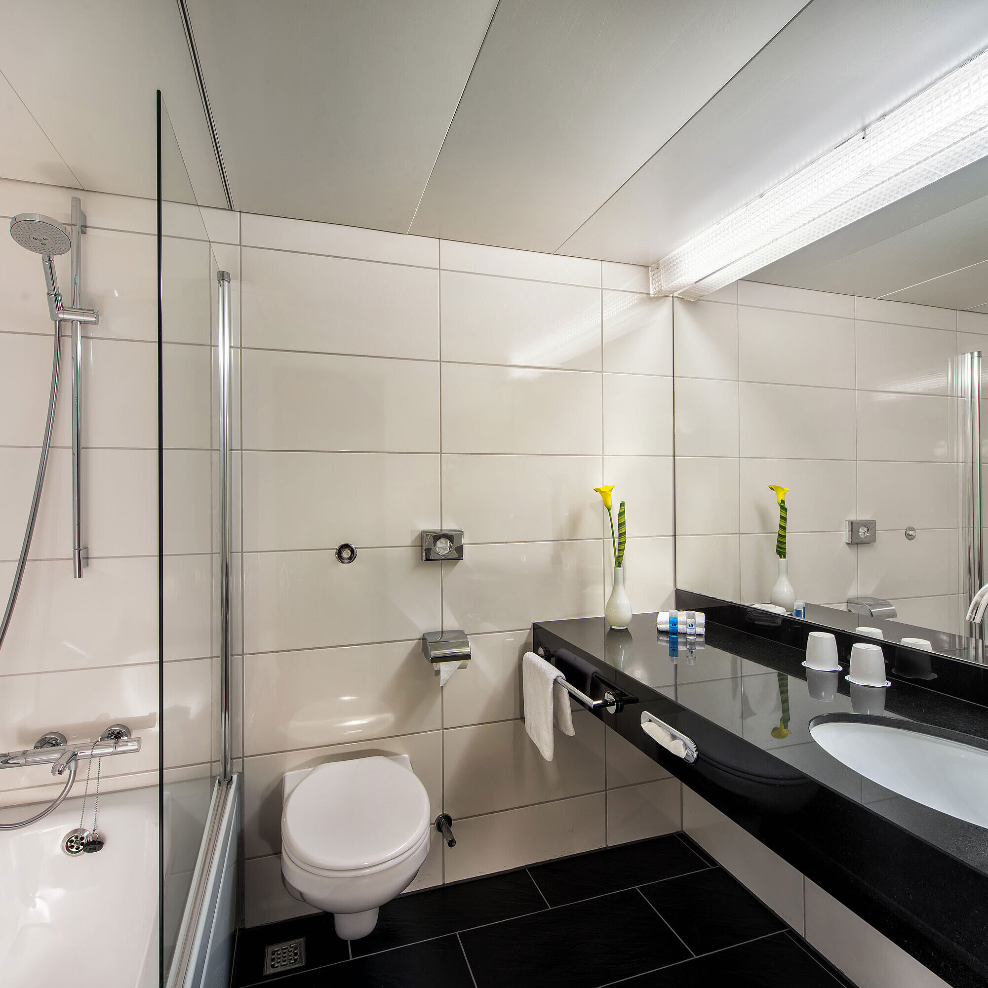 Baño Cuarto de baño del Maritim Hotel Kiel con bañera, mampara de ducha de cristal, lavabo y gran espejo sobre repisa negra.