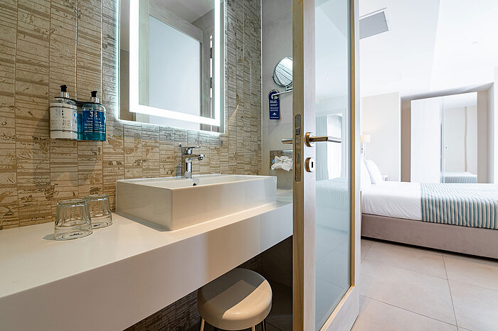 Suite Junior Moderno bagno della Junior Suite del Maritim Antonine Hotel & Spa Malta con specchio illuminato, elegante lavabo e vista sulla luminosa zona notte.