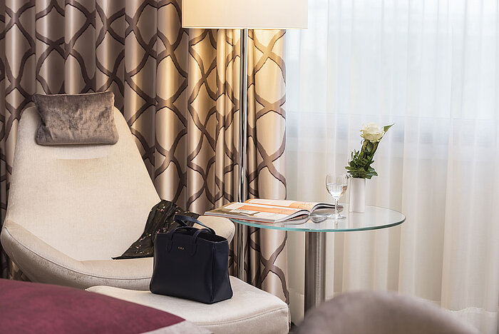 Detalle de una habitación Classic en el Hotel Maritim Bonn con silla cómoda, cortina, mesa, vaso, rosa y bolso. 