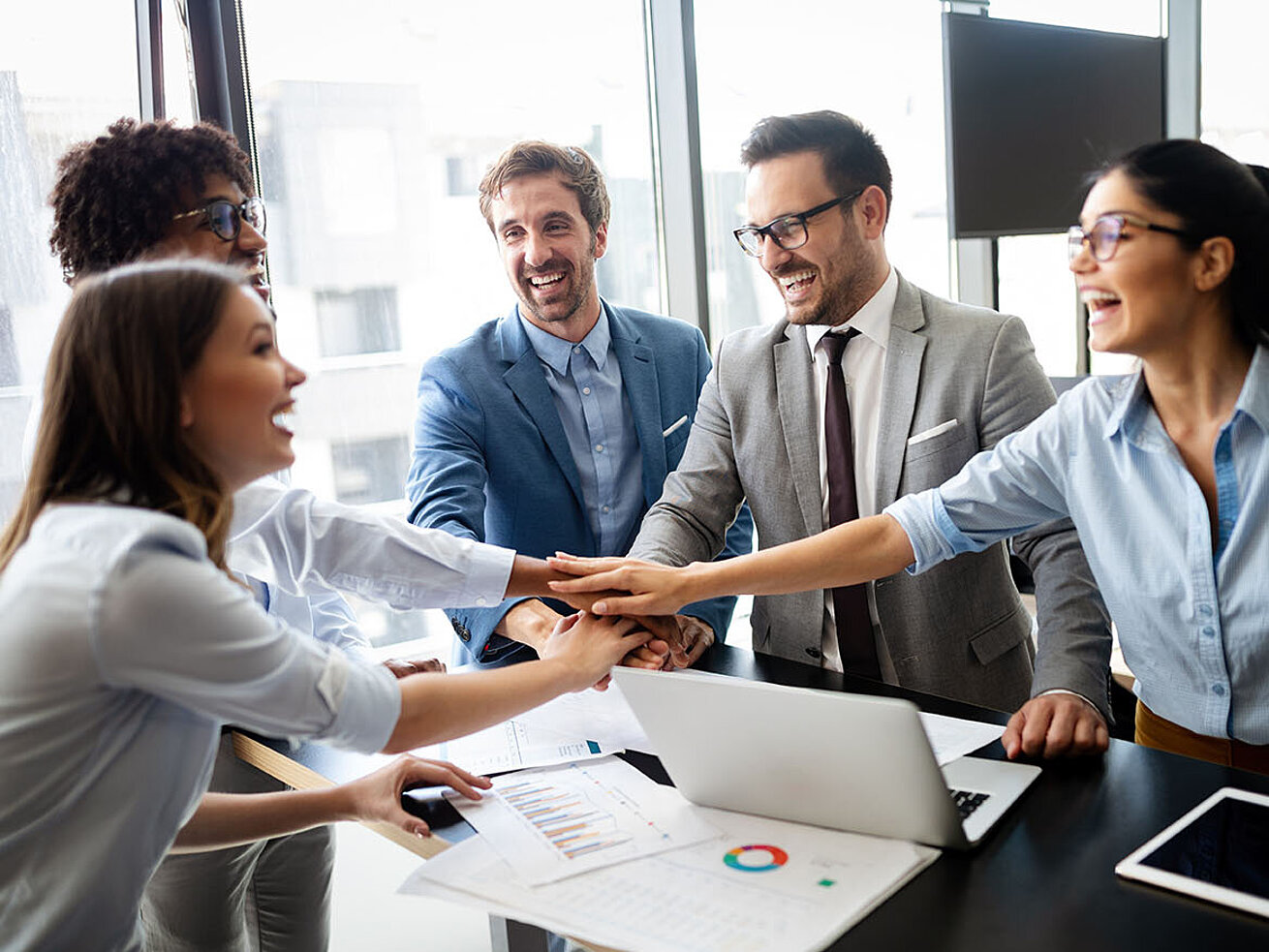 © NDABCREATIVITY - AdobeStock.com Motivated business team in office joining hands during meeting with laptop and documents on conference table