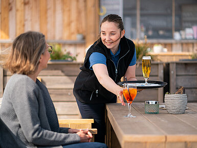 Una cameriera serve Aperol Spritz e birra a un ospite sulla terrazza del Beach Lounge, con la spiaggia sullo sfondo.