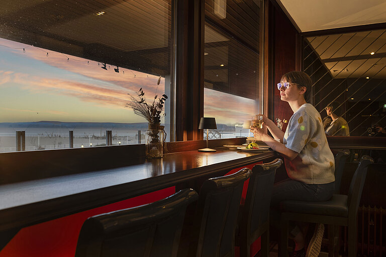 Huésped con bebida junto a la ventana viendo el atardecer en el Maritim Hotel Travemünde