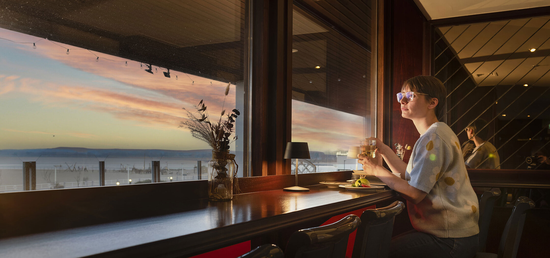 Huésped con bebida junto a la ventana viendo el atardecer en el Maritim Hotel Travemünde