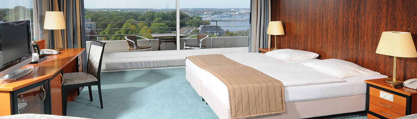 Spacious hotel room with a double bed, seating area and wide views of Kiel through the large window front.