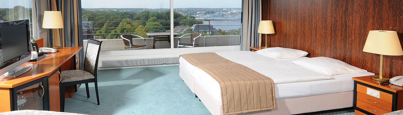 Camera Comfort Ampia camera d’albergo con letto matrimoniale, area salotto e vista su Kiel attraverso la grande vetrata.