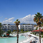 Panorama del Maritim Resort Calabria con piscina, palmeras, edificios blancos y montañas al fondo bajo un cielo despejado.