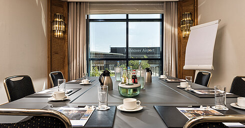 Sala de reuniones luminosa con mesa de conferencias, bebidas y vista al aeropuerto en el Maritim Hotel Hannover.