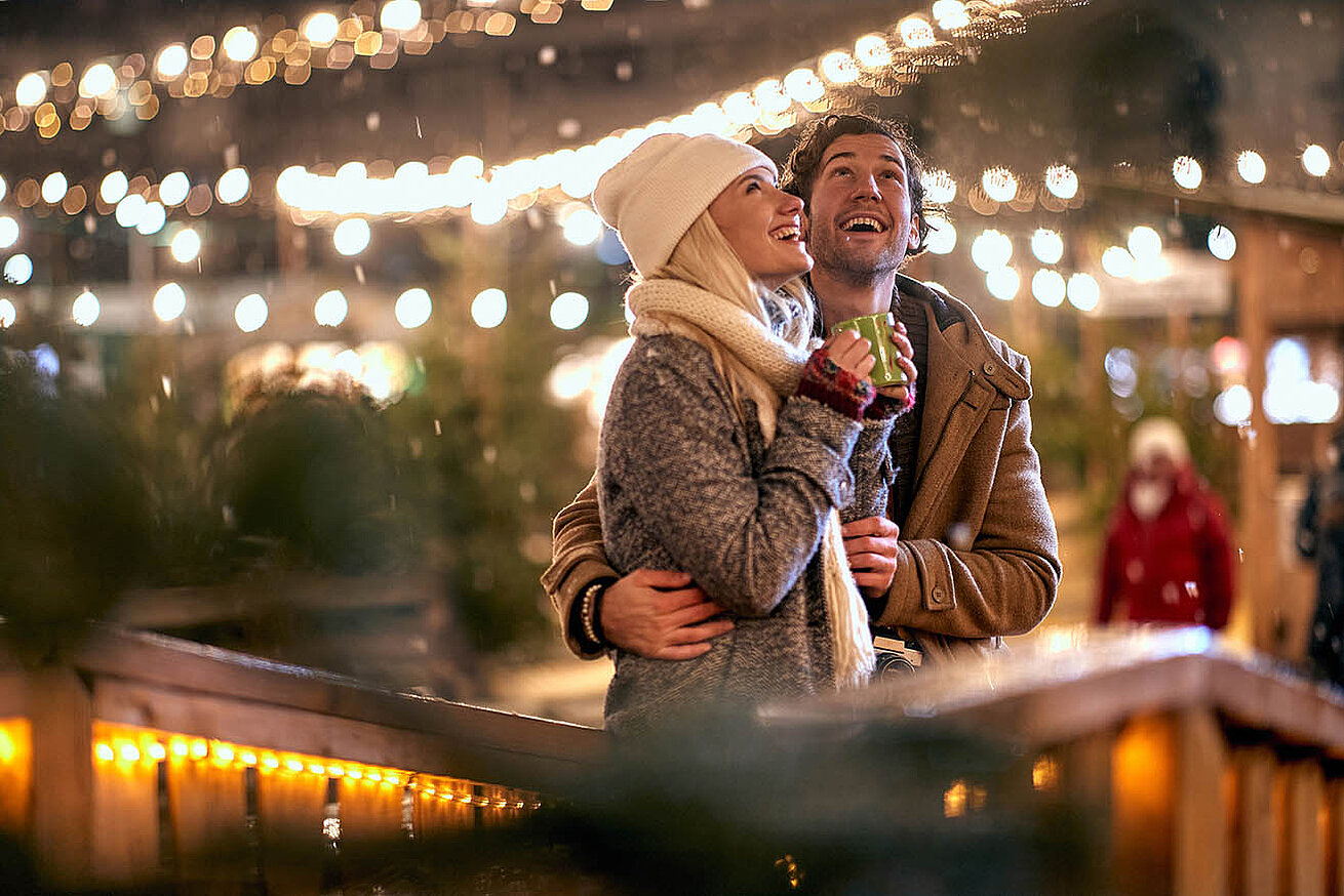 © luckybusiness - AdobeStock.com Coppia che si gode una serata invernale al mercatino di Natale, tra luci calde e atmosfera festosa.