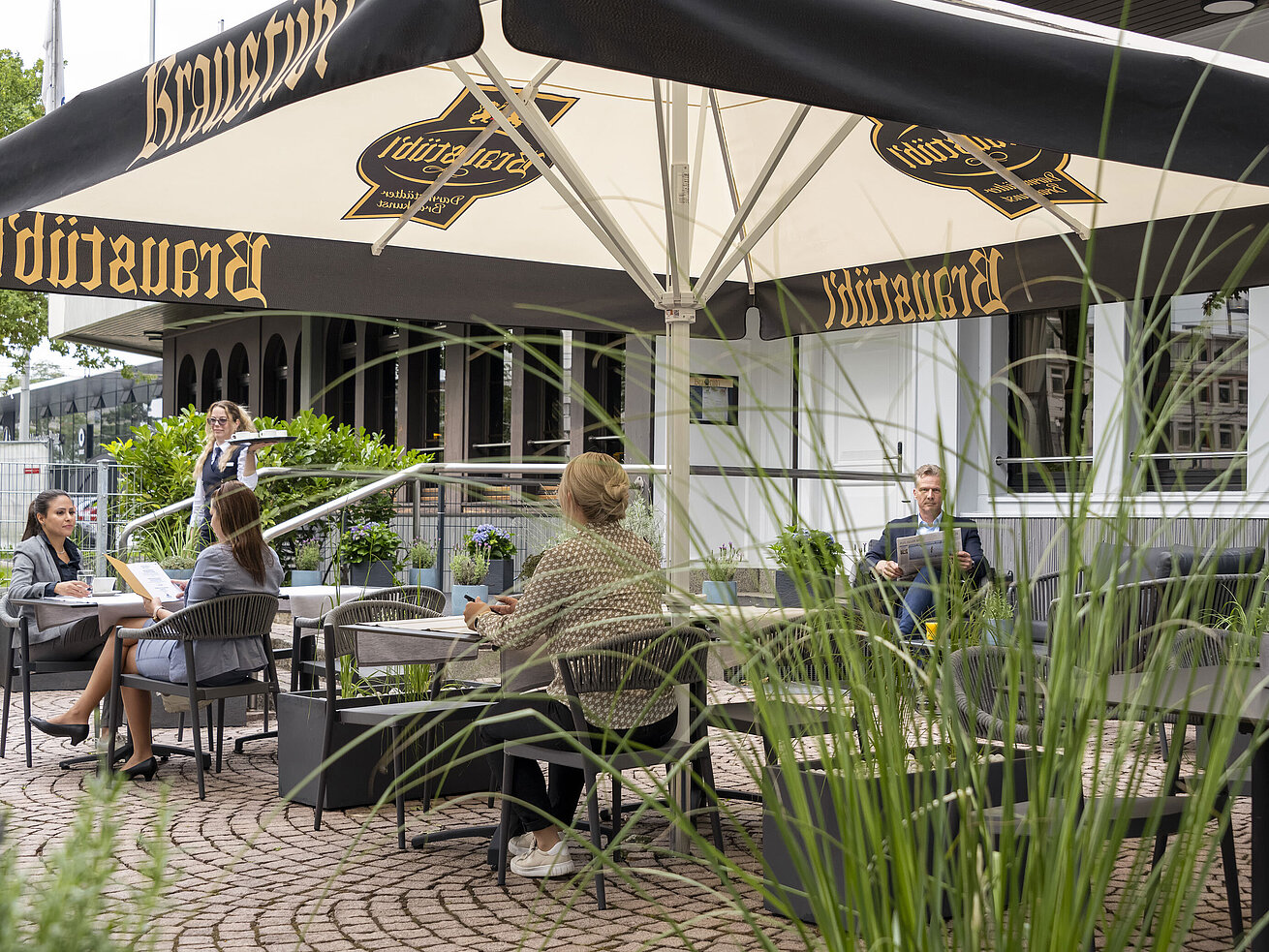 Terrace Guests enjoying a meal on the terrace of the Maritim Hotel Darmstadt under large umbrellas with Brauhaus logo.