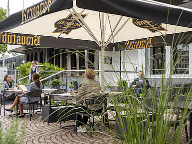 Guests enjoying a meal on the terrace of the Maritim Hotel Darmstadt under large umbrellas with Brauhaus logo.
