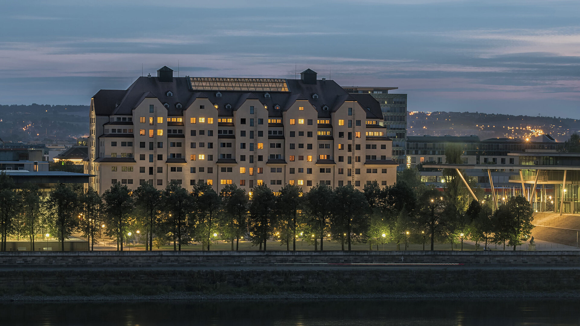 Illuminated Maritim Hotel Dresden at dusk with lights reflecting on the Elbe River.