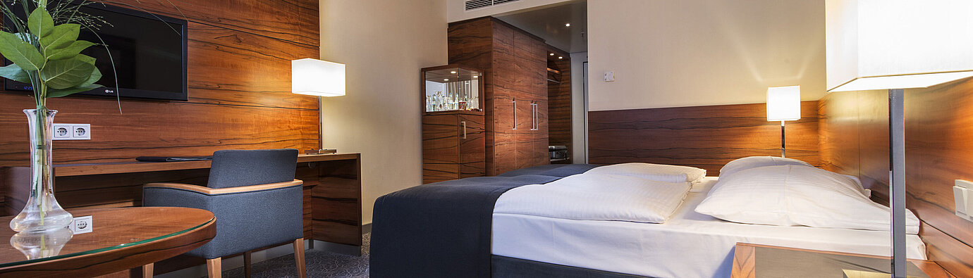 Habitación de hotel amueblada en el Maritim Hotel Múnich con cama cómoda, zona de trabajo, TV y paneles de madera.