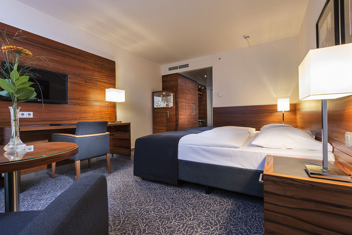 Habitación de hotel amueblada en el Maritim Hotel Múnich con cama cómoda, zona de trabajo, TV y paneles de madera.