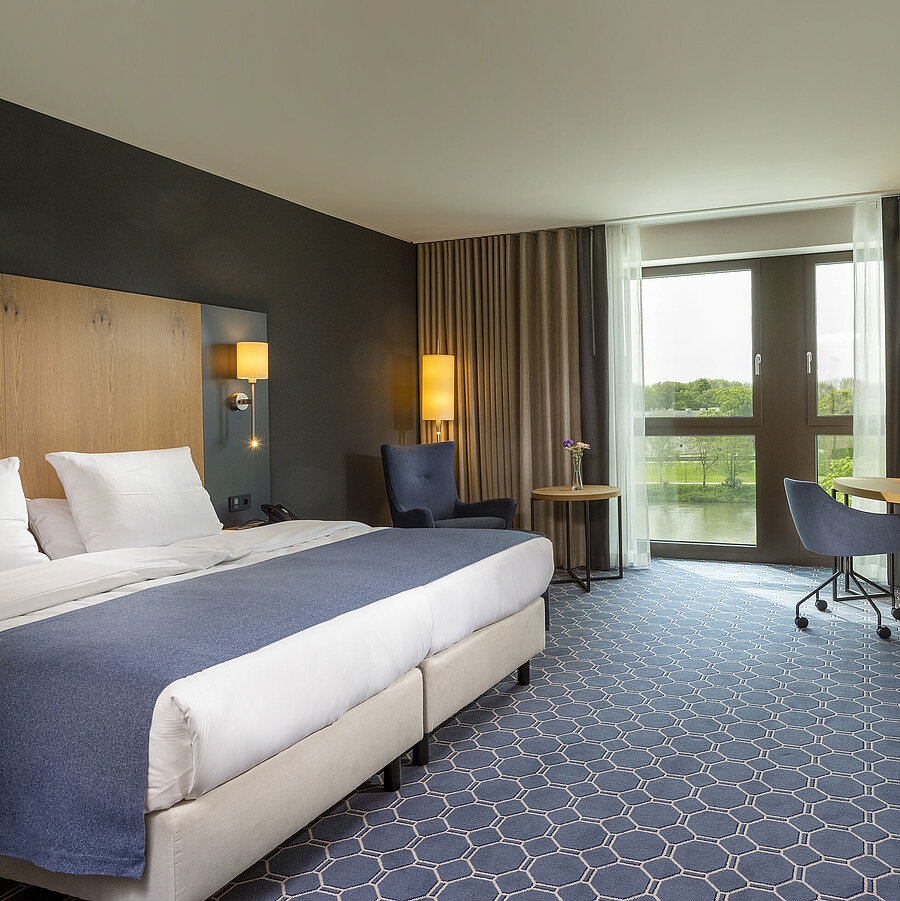 Chambre double spacieuse à l’hôtel Maritim Ingolstadt avec grand lit, bureau et vue sur la rivière.