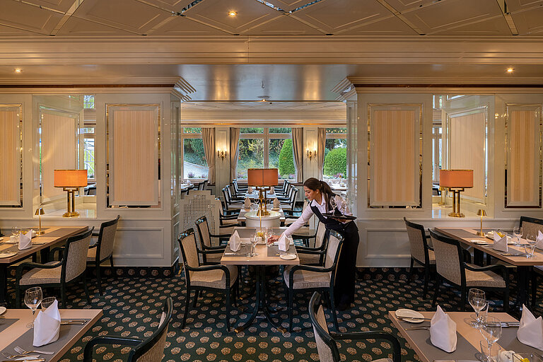 Restaurant Maritim Restaurant élégant avec tables dressées, grandes fenêtres et membre du personnel préparant la salle.