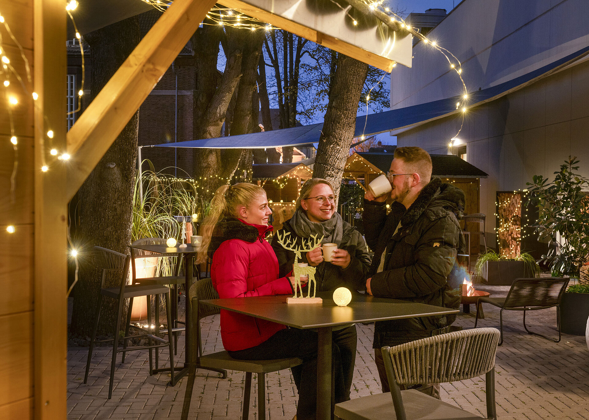 Maritim Christmas Village Three people enjoying hot drinks at the festive Christmas market of Maritim Hotel Darmstadt with fairy lights.