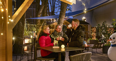Three people enjoying hot drinks at the festive Christmas market of Maritim Hotel Darmstadt with fairy lights.