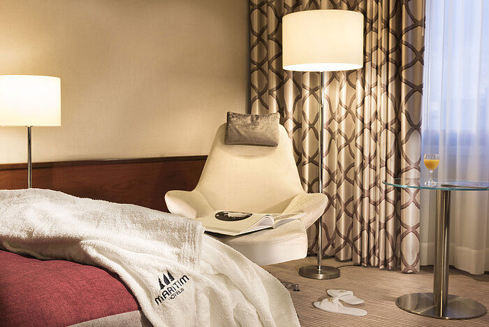 Detalle de una habitación Comfort en el Maritim Hotel Bonn con sillón, mesa, vaso de zumo y albornoz en la cama.