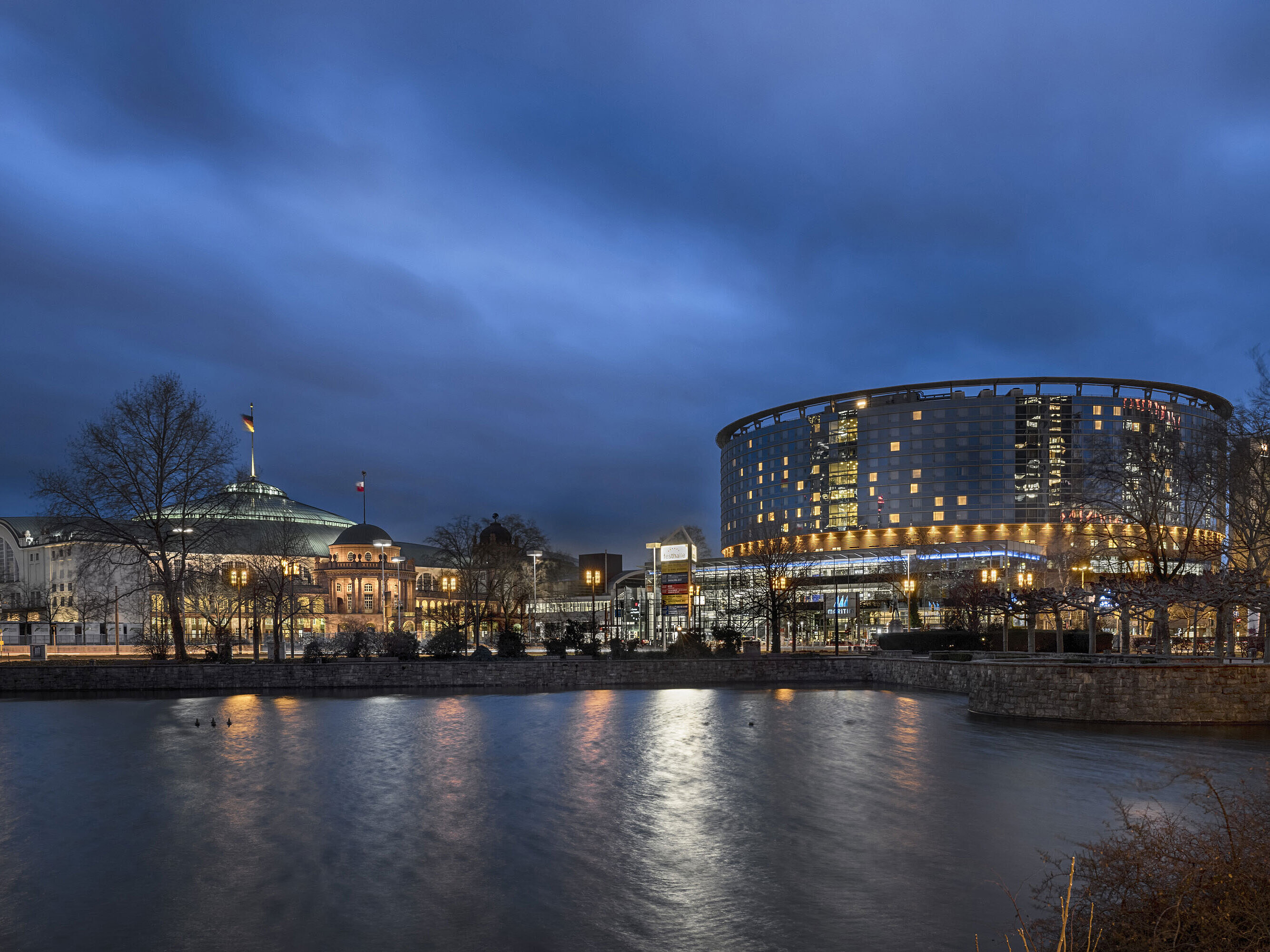 Hotel Maritim Frankfurt e Festhalle di notte con facciata in vetro illuminata e riflesso nello stagno adiacente.