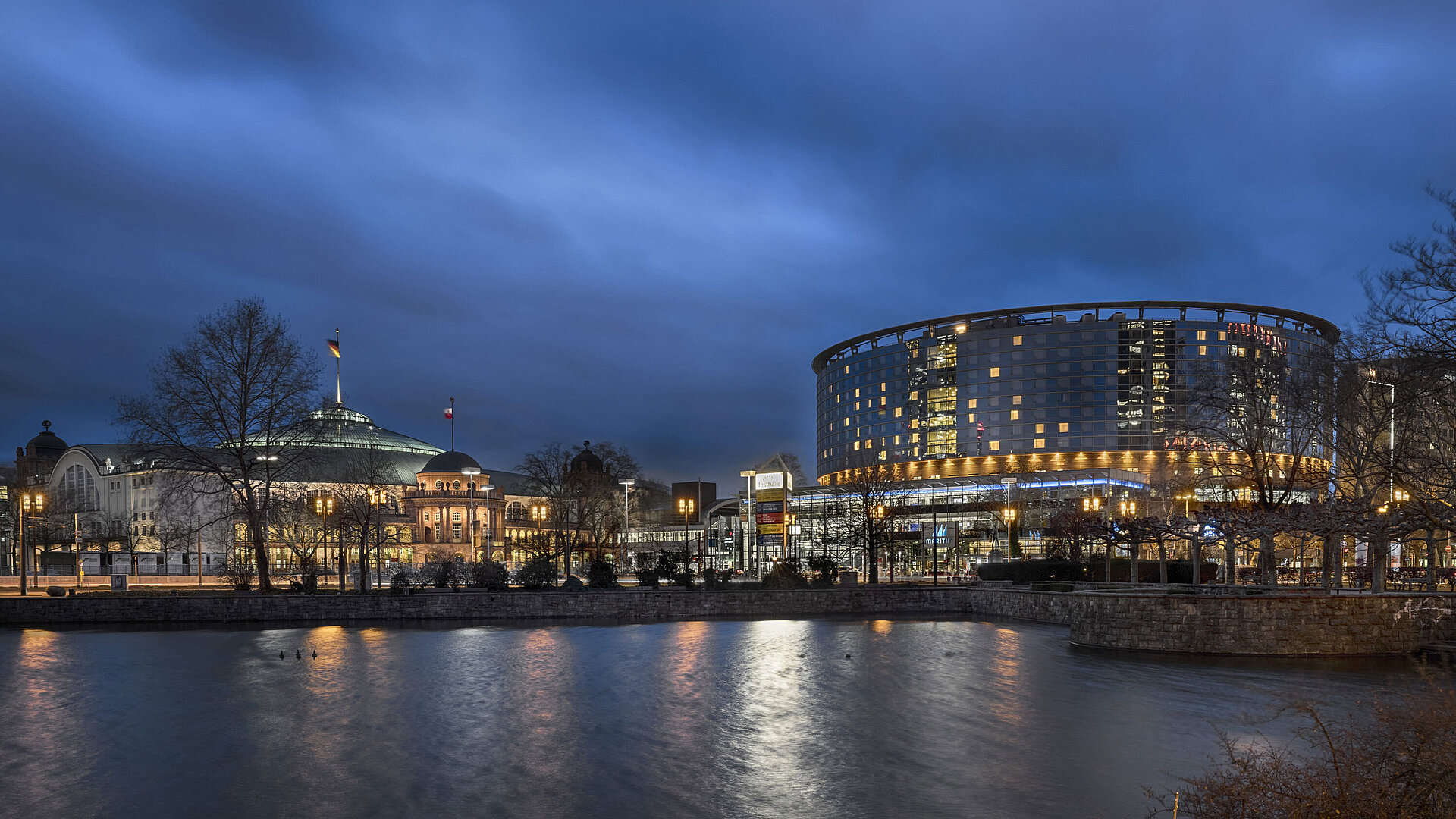 Maritim Hotel Frankfurt and Festhalle at night with illuminated glass facade and reflection in the adjacent pond.