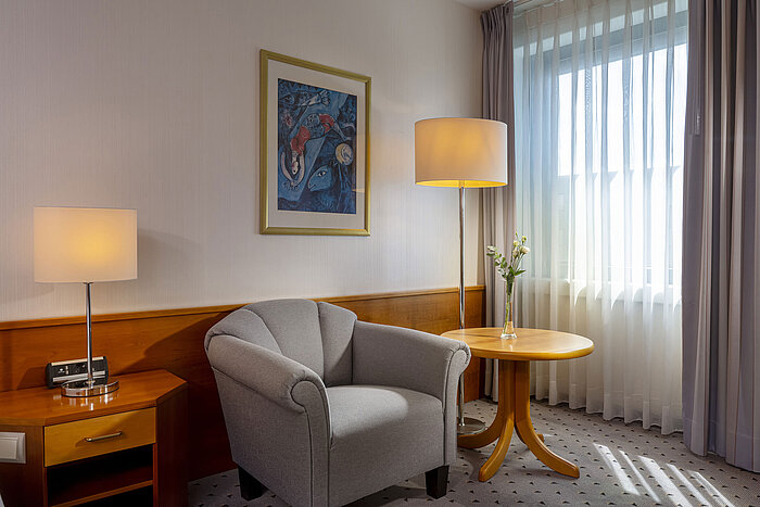 Habitación de hotel luminosa en el Maritim Airport Hotel Hannover con sillón, lámpara, ventana y obra de arte.