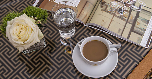 Primer plano con rosa, café, agua y revista sobre mesa de cristal