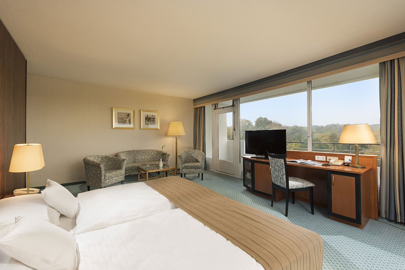 Chambre Comfort du Maritim Hotel Kiel avec un coin salon confortable, un bureau, un grand lit et une vue magnifique sur la verdure.
