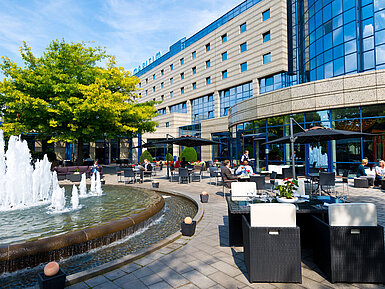 Terraza del Hotel Maritim Bonn con áreas de descanso, fuente y huéspedes cenando y relajándose al aire libre.
