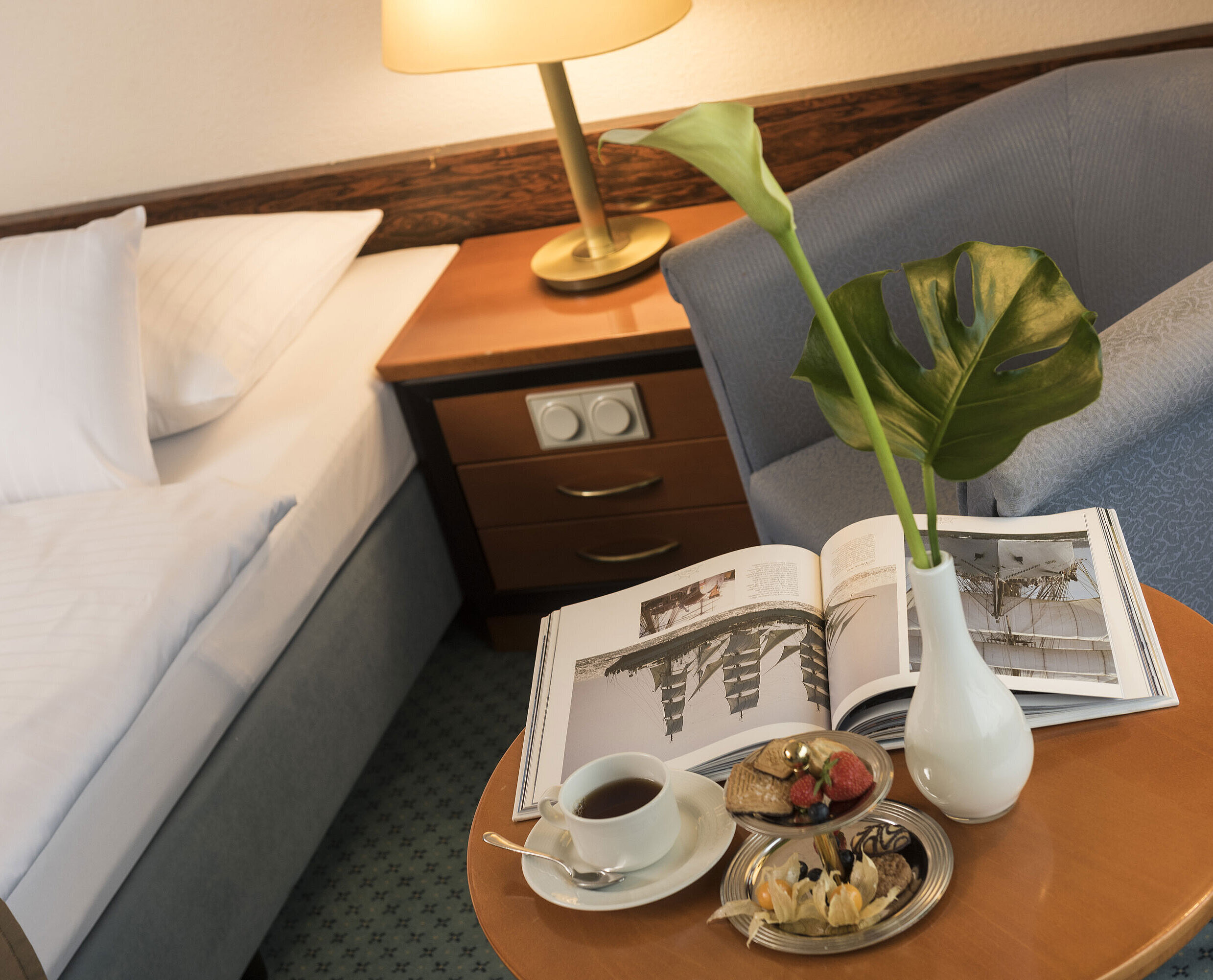 Detalle de la sala Detalles de una habitación del Maritim Hotel Kiel: mesa auxiliar con jarrón, libro y aperitivos junto a una cama y un sillón.
