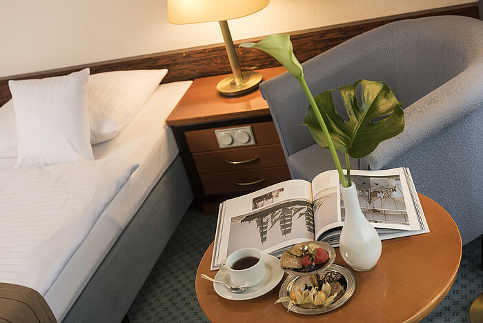 Détails d'une chambre du Maritim Hotel Kiel : table d'appoint avec vase, livre et snacks à côté d'un lit et d'un fauteuil.