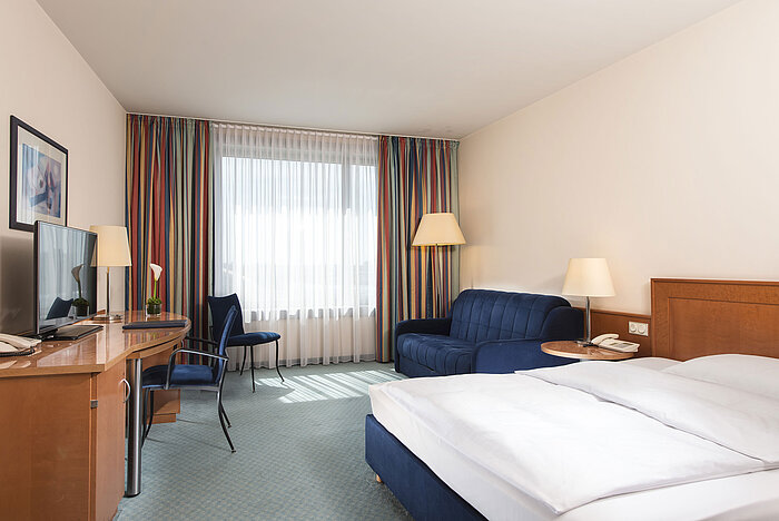 Chambre classique à l'hôtel Maritim Frankfurt avec lit double, bureau, canapé et grandes fenêtres pour une lumière naturelle.