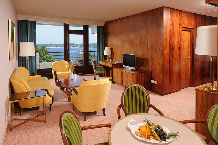 Junior Suite au Maritim Hotel Kiel, coin salon confortable, bureau, TV et balcon avec vue sur l'eau.