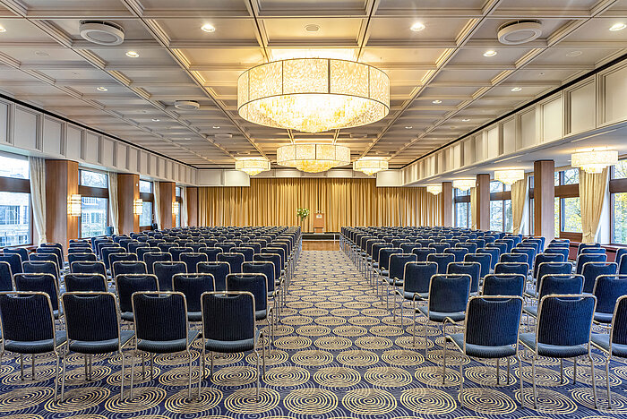 Saal Maritim nel Maritim Hotel Kiel, grande sala conferenze con posti a sedere in fila, lampadari e un palco davanti a un sipario.