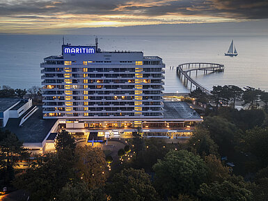 Vue du soir du Maritim Seehotel Timmendorfer Strand avec jetée illuminée et voilier en arrière-plan au coucher du soleil.