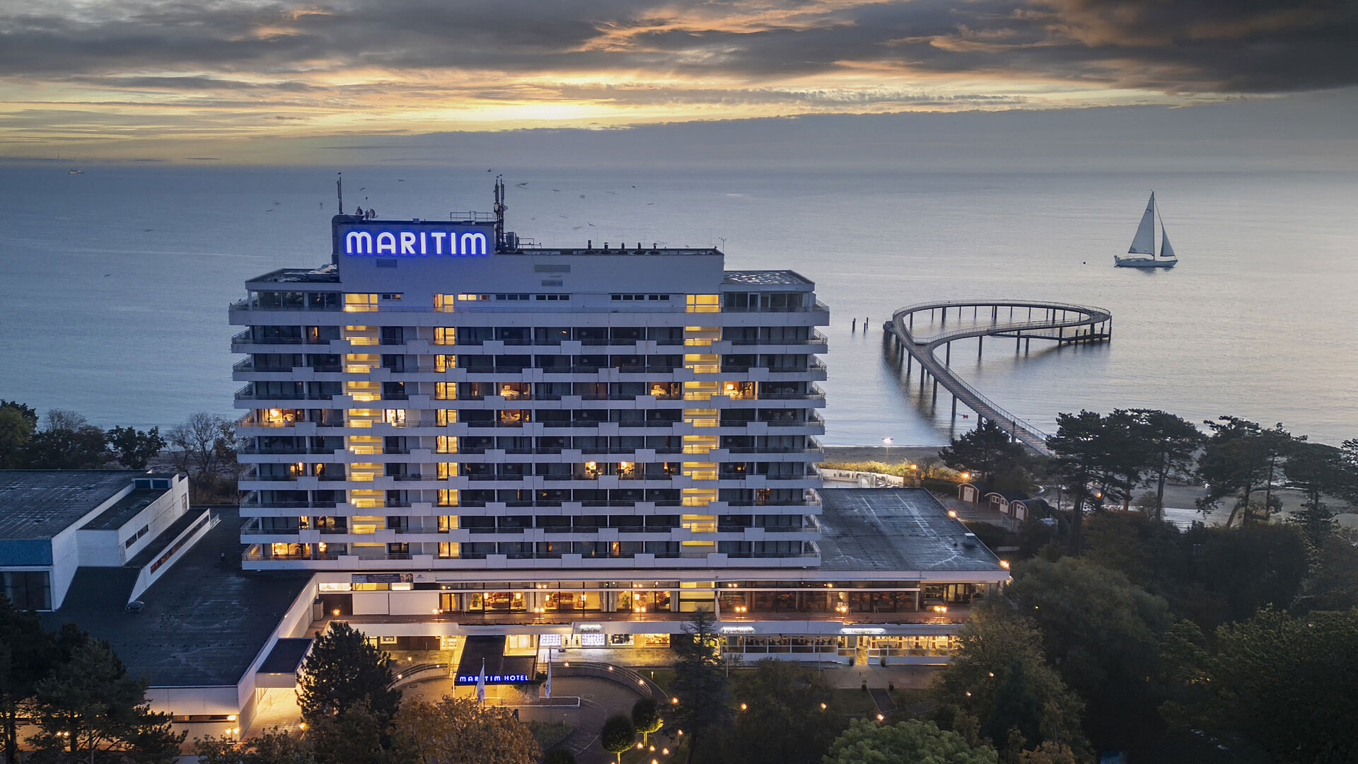 Evening view of Maritim Seehotel Timmendorfer Strand with illuminated pier and a sailboat in the background at sunset.