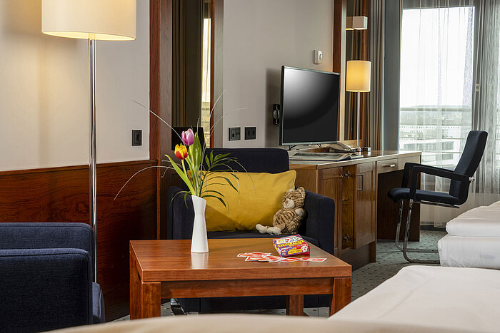 Habitación familiar en el Maritim Darmstadt con sillón acogedor, mesa con cartas y jarrón de tulipanes, y escritorio con TV.