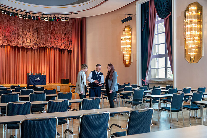 Waldeck Hall at Maritim Hotel Bad Wildungen with row seating and three people talking in front of the stage.