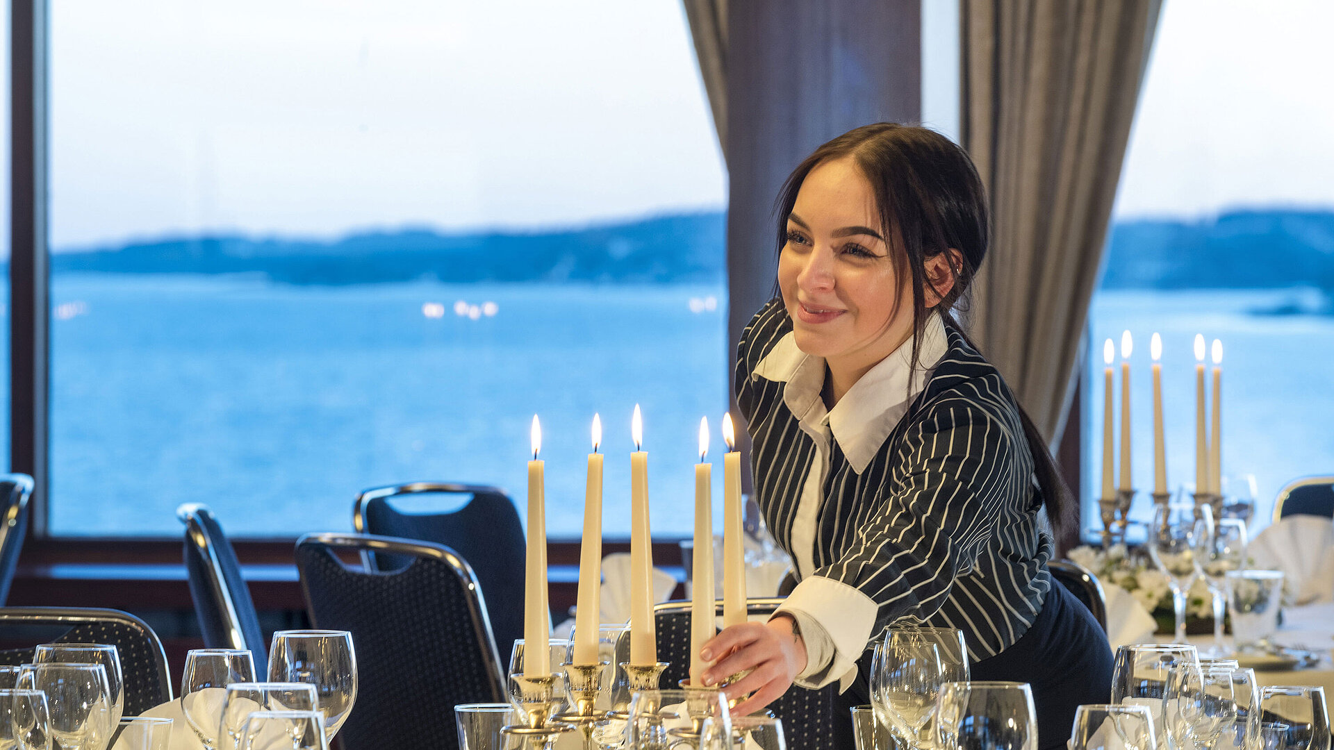Hall Maritim en el Hotel Maritim de Kiel, un miembro del personal enciende velas en una mesa festiva, con vistas al mar al fondo.