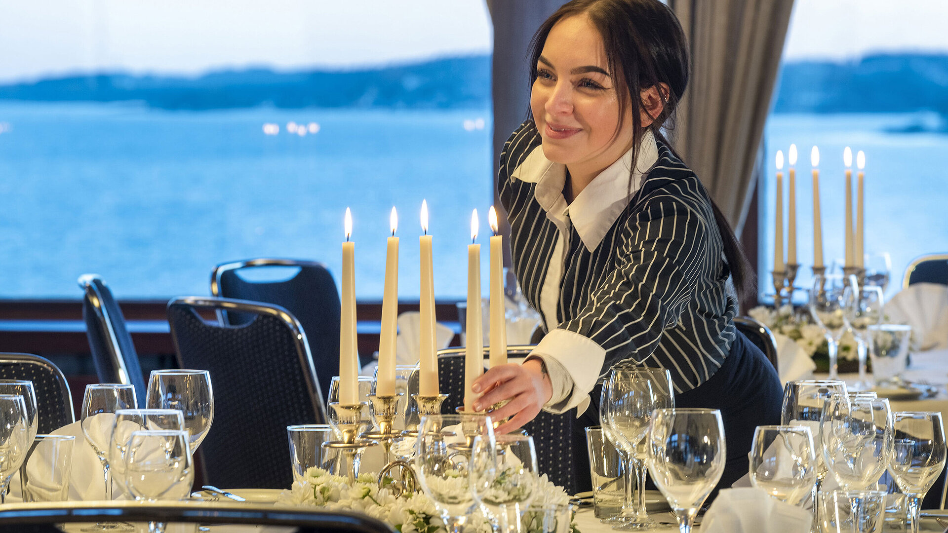Hall Maritim en el Hotel Maritim de Kiel, un miembro del personal enciende velas en una mesa festiva, con vistas al mar al fondo.