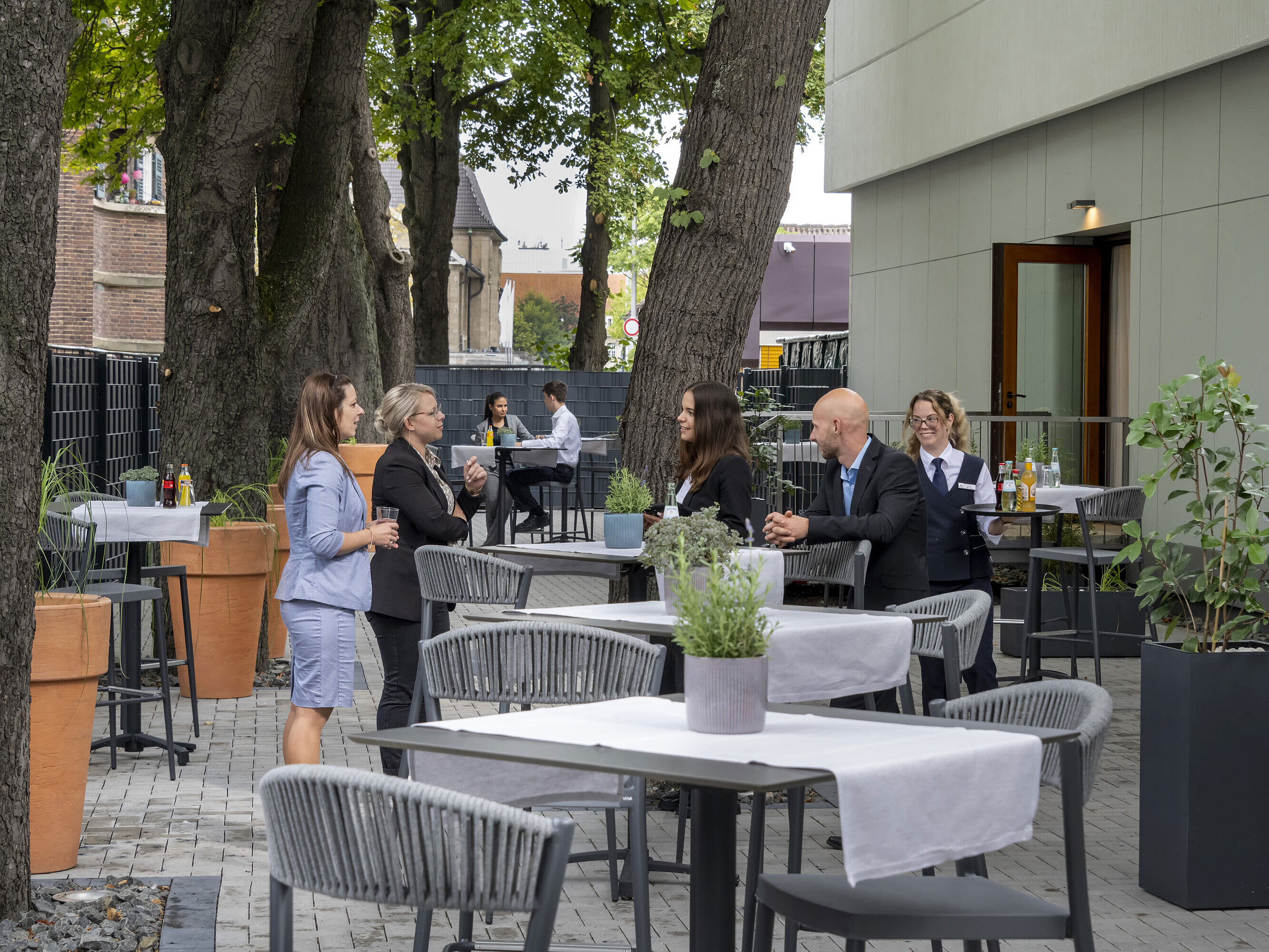 Terraza moderna del hotel Maritim Darmstadt con invitados relajándose, árboles al fondo y elegantes muebles grises.