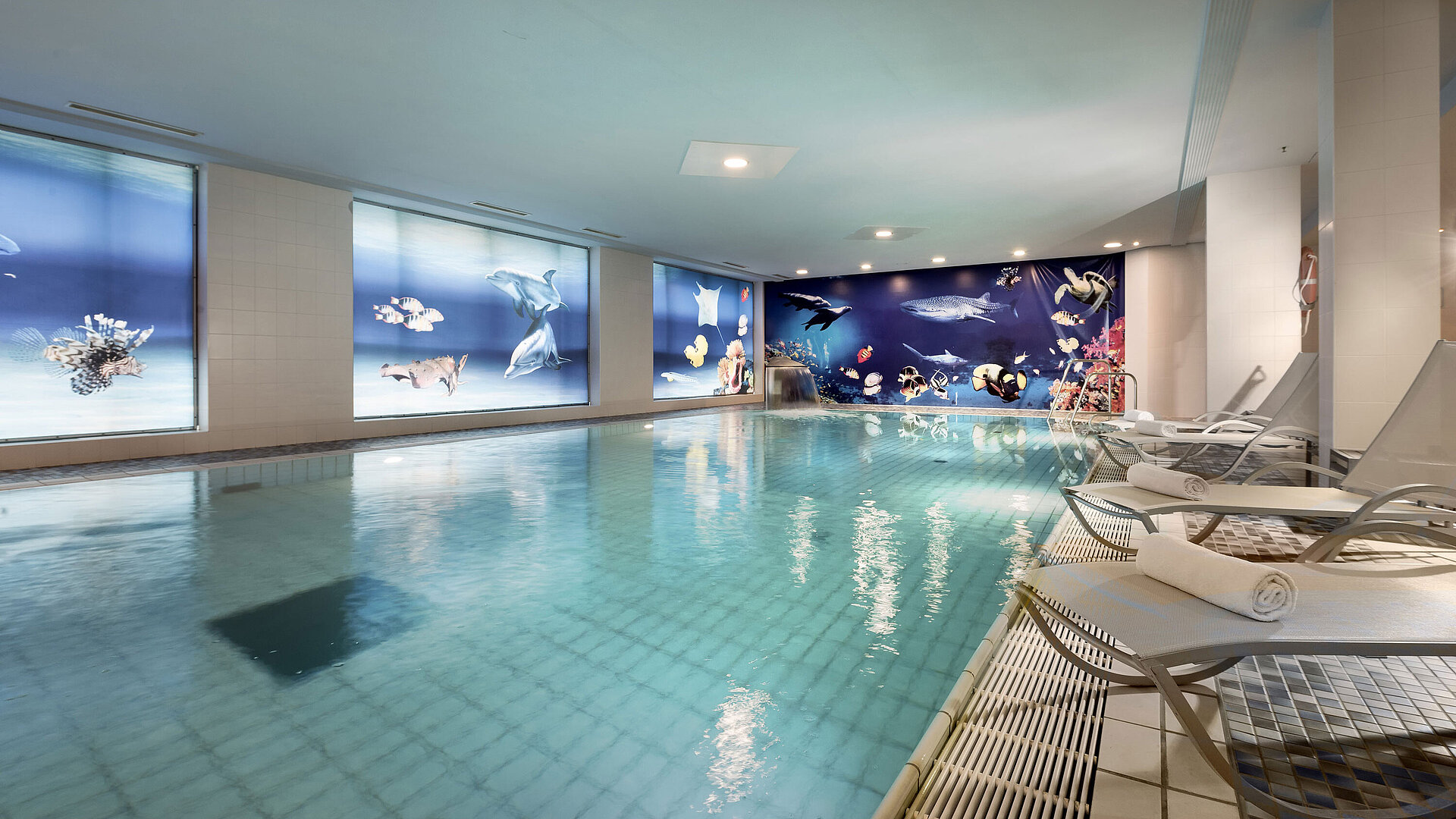 Piscina coperta del Maritim proArte Hotel Berlin con lettini rilassanti e straordinarie decorazioni subacquee sulle pareti.