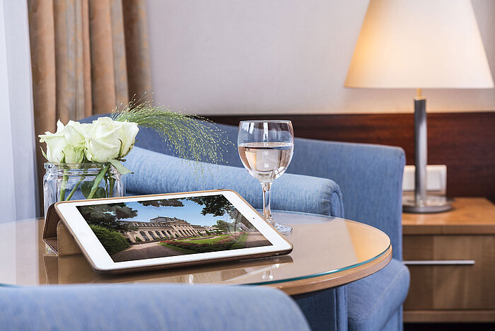 Accogliente area relax al Maritim Hotel Fulda con tablet, bicchiere d’acqua e rose bianche su tavolo in vetro.