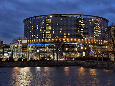 Hotel Maritim Frankfurt di notte con facciata in vetro illuminata e riflesso nello stagno tranquillo.