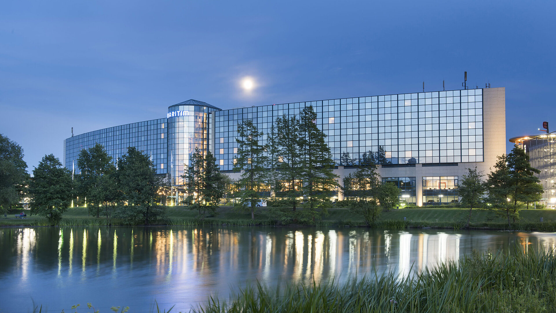 Illuminated Maritim Hotel Hannover with a glass facade and lake reflection at dusk.