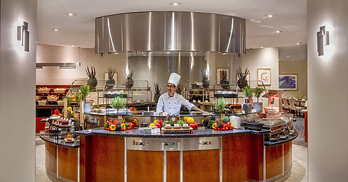 Chef alla stazione di show-cooking del Maritim proArte Hotel Berlin, circondato da verdure e erbe fresche.